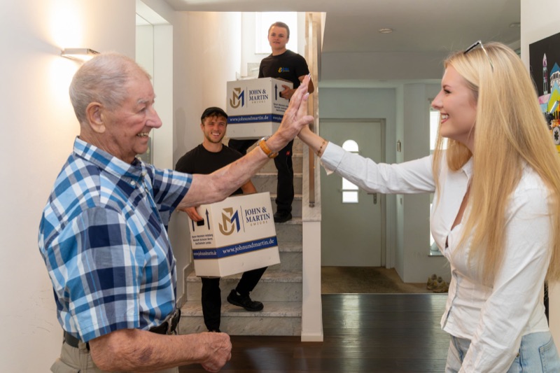 Im neuen Zuhause klatschen sich ein Kunde und eine junge Frau ab, im Hintergrund tragen Mitarbeiter Kartons.