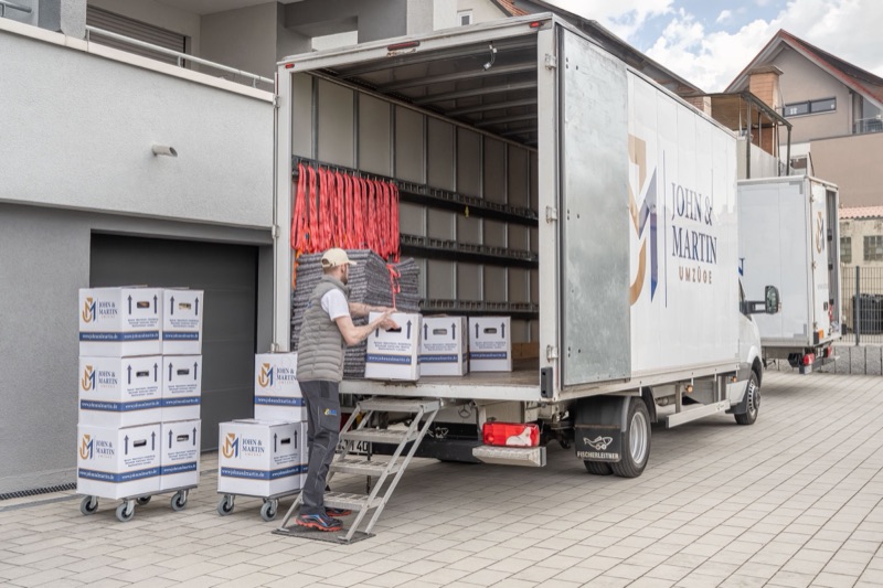 Ein Mitarbeiter von John & Martin belädt einen Umzugswagen mit Firmenlogo, daneben stehen Umzugskartons auf Rollwagen.
