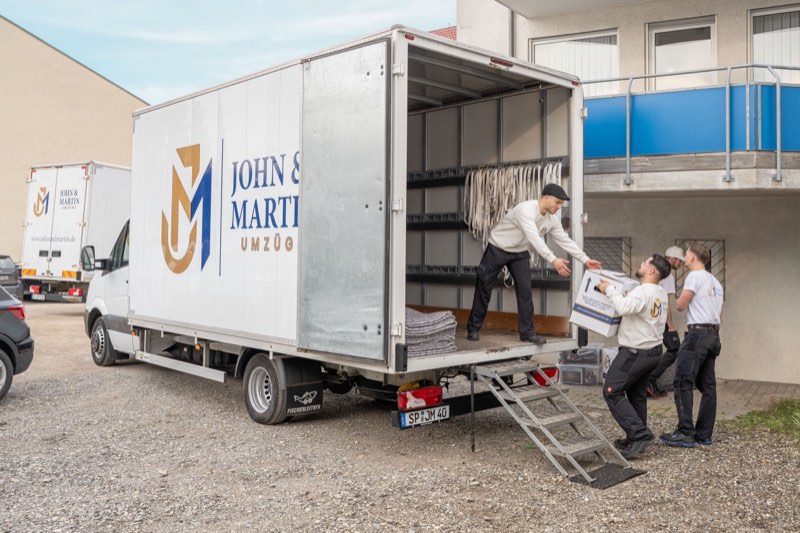 Drei Mitarbeiter von John & Martin reichen Kartons über eine Rampe in einen Umzugswagen mit Firmenlogo.
