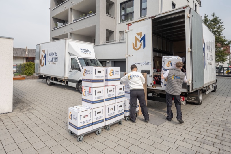 Mitarbeiter beladen zwei Umzugswagen von John & Martin, davor stehen Kartons auf Rollwagen.