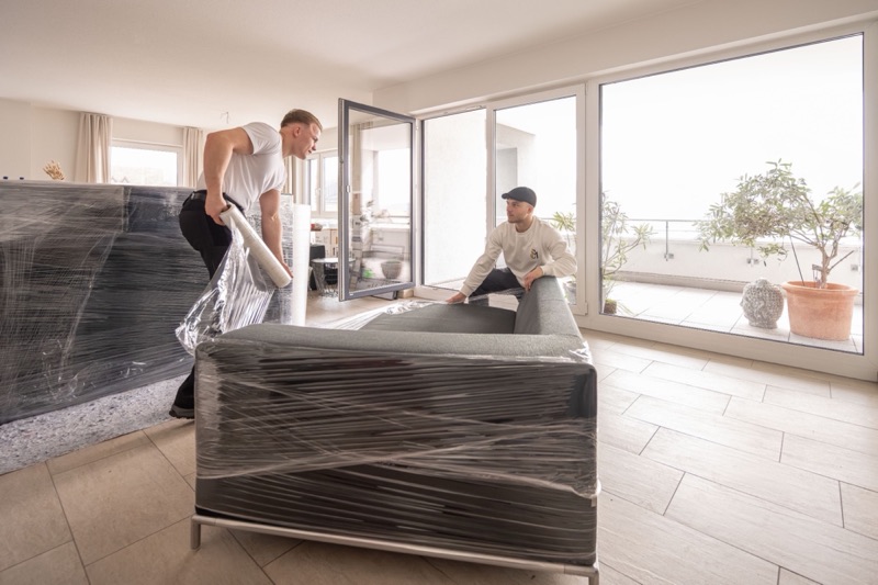 Zwei Mitarbeiter wickeln in einer hellen Wohnung ein Sofa in Stretchfolie.