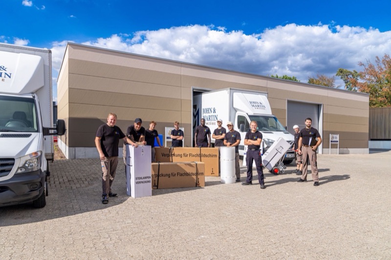 Das Team von John & Martin vor der eigenen Halle mit zwei Umzugswagen, daneben Schutzkartons für Flachbildschirme und Stehlampen.