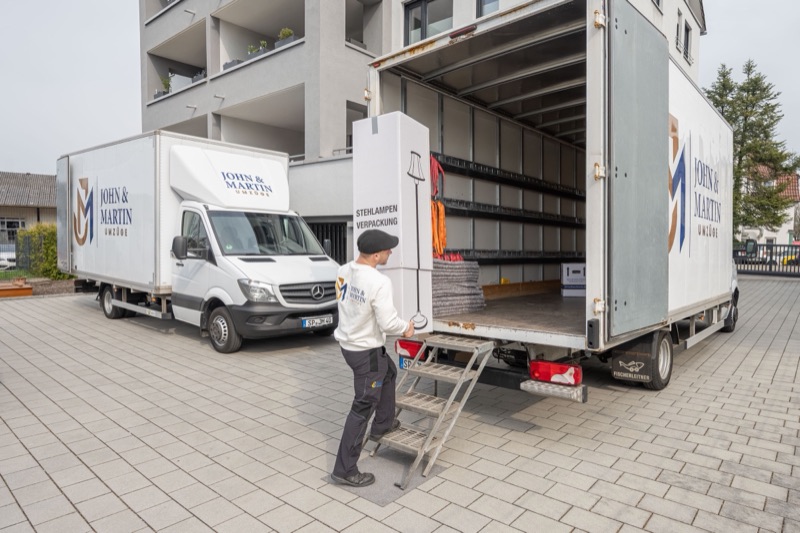 Ein Mitarbeiter von John & Martin schiebt einen Schrankkarton über die Ladebordwand in den Umzugswagen, daneben steht ein zweiter Wagen.