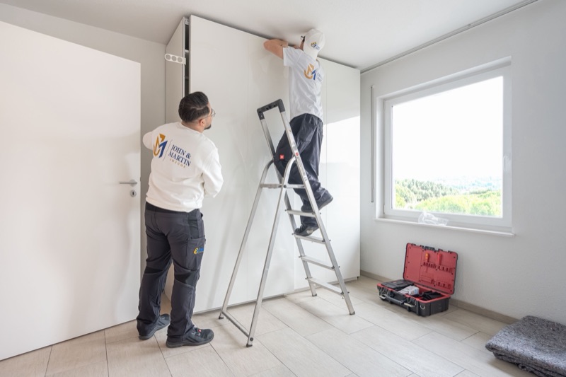 Zwei Mitarbeiter von John & Martin bauen einen Kleiderschrank auf, einer steht auf der Leiter.