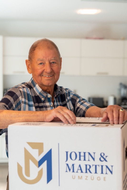 Ein älterer Herr lächelt in seiner neuen Küche, vor ihm ein Karton von John & Martin.