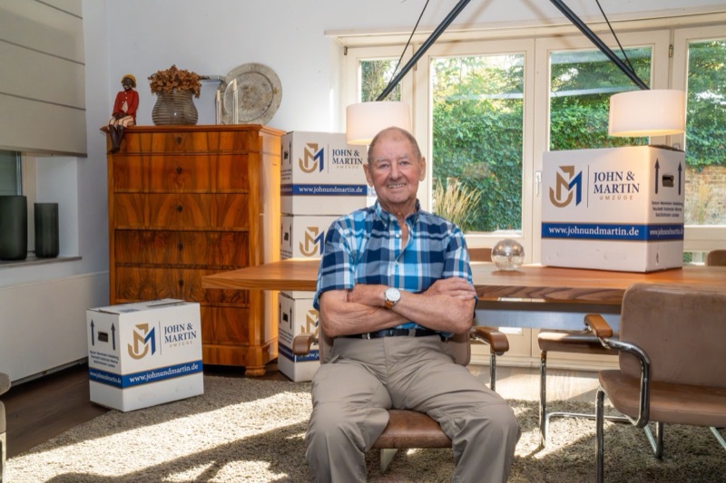 Ein älterer Herr sitzt zufrieden in seinem Esszimmer zwischen Umzugskartons von John & Martin.
