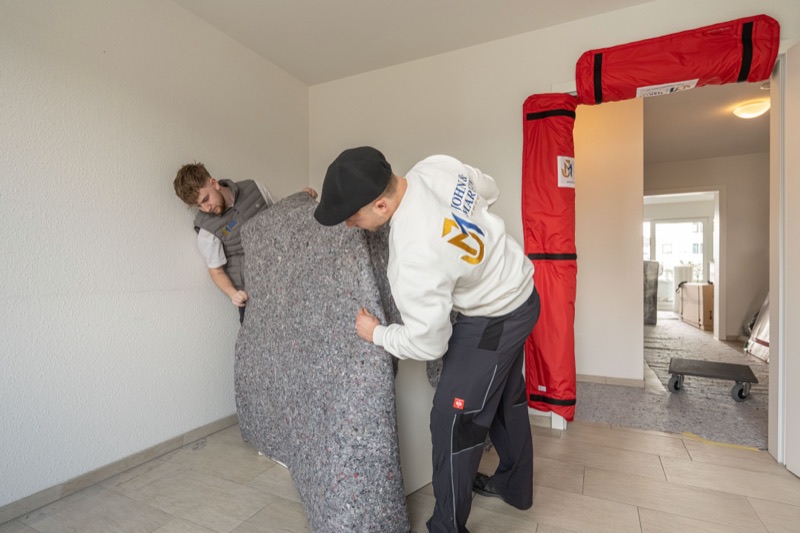 Zwei Mitarbeiter hüllen ein Möbelstück in eine graue Umzugsdecke.