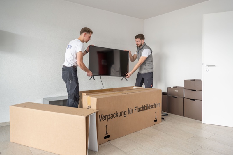 Zwei Mitarbeiter heben einen Fernseher in einen Karton für Flachbildschirme.
